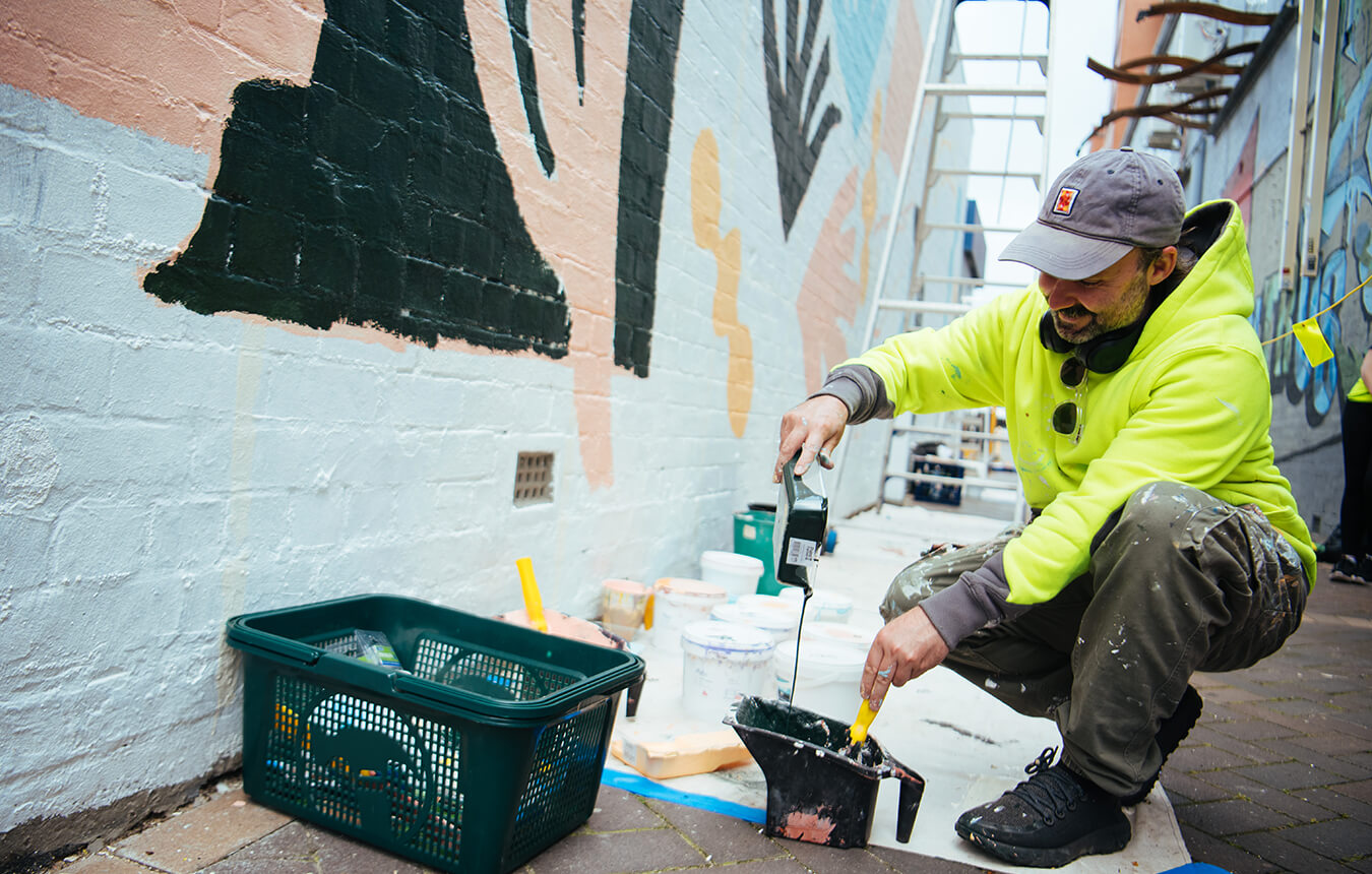 Sean Thomas Bell pouring paint into container in front of mural in Union Lane, Newcastle