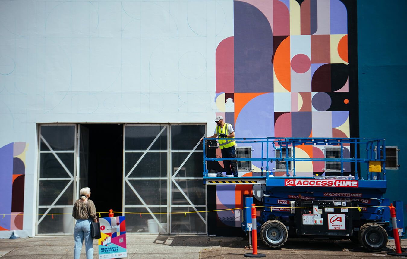 Taj 'Deams' Alexander in scissor lift talking to a woman in front of mural in Parry Street, Newcastle